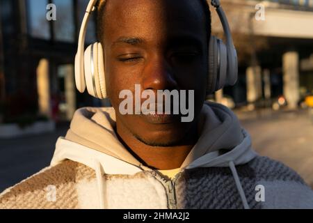 Thoughtful African American male listening to music in wireless headphones while standing with eyes closed on sunny street near modern building in cit Stock Photo