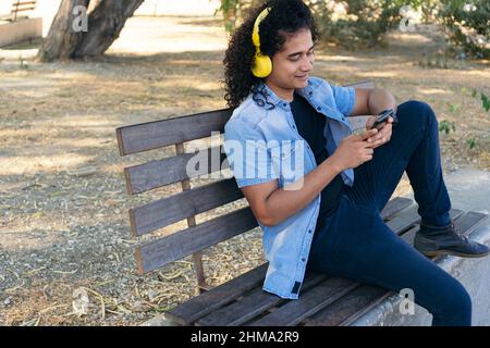 Young smiling Hispanic male in casual clothes listening to music in wireless headphones and browsing cellphone on bench in park Stock Photo
