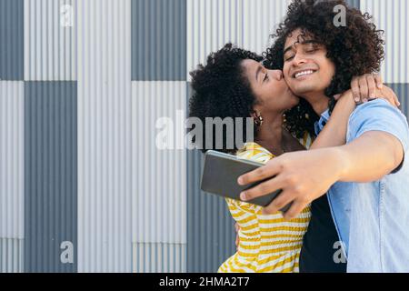 Happy curly haired women embracing smiling Hispanic male partner taking self portrait on cellphone against metal ribbed wall Stock Photo