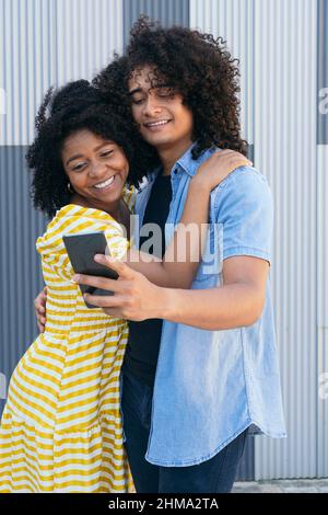 Happy curly haired women embracing smiling Hispanic male partner taking self portrait on cellphone against metal ribbed wall Stock Photo