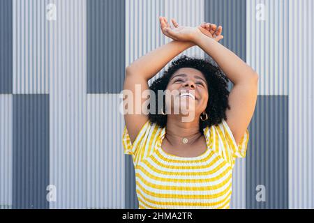 Happy Hispanic female with curly hair standing near metal background looking up Stock Photo