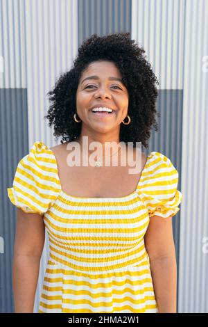Happy Hispanic female with curly hair standing near metal background looking at camera Stock Photo