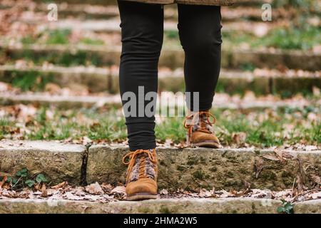 Crop anonymous person in black trousers and sneakers walking down shabby stone steps with fallen leaves on autumn day in park Stock Photo