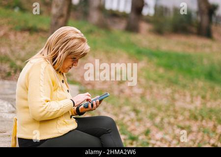 Side view middle aged female text messaging on modern cellphone while sitting in park with trees on blurred background Stock Photo