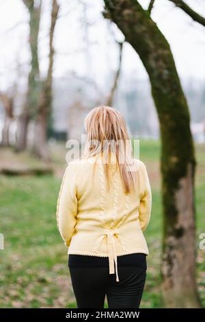 Back view middle aged Woman standing in park with tall green trees on blurred background Stock Photo