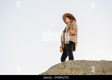 From below of female tourist in hat standing on rocky formation and looking away under cloudless sky in sunny day Stock Photo