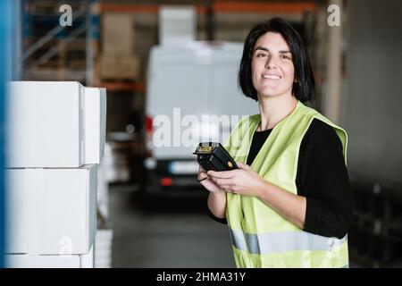 Side view of smiling female warehouse worker in uniform high vis vest looking at camera while using professional portable data terminal checking suppl Stock Photo