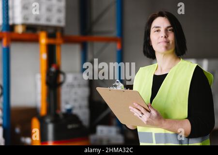 Warehouse female worker in uniform high vis vest writing notes on clipboard while checking supplies during work in storehouse looking at camera Stock Photo