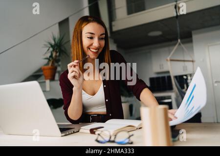 Happy young red haired female freelancer sitting at table with netbook while analyzing diagrams and taking notes in notebook and working remotely at h Stock Photo