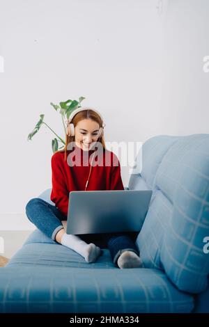 Full body of cheerful young red haired female resting on sofa with netbook while listening to music in headphones and enjoying weekend at home looking Stock Photo