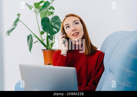 Thoughtful young female with red hair resting on sofa with netbook while speaking on smartphone in light living room at home Stock Photo