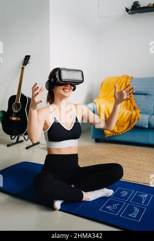 Side view full length of anonymous smiling female in sportswear on mat siting with crossed legs while training in VR goggles at home Stock Photo