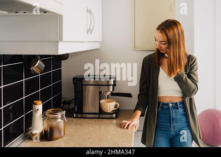 Young red haired female making coffee in machine while talking on smartphone in kitchen at home in morning and looking down Stock Photo