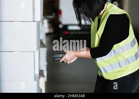 Side view of crop anonymous female warehouse worker in green uniform high vis vest scanning barcode on box with special portable data terminal Stock Photo