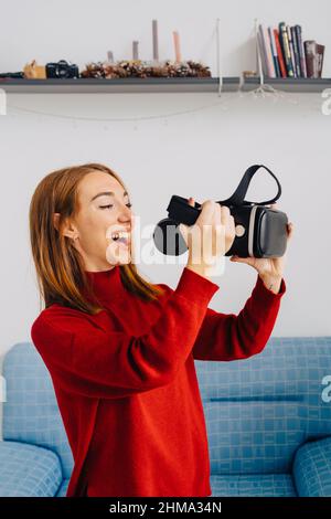 Cheerful Woman holding VR goggles standing against sofa and exploring virtual reality Stock Photo
