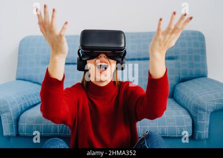 Woman in VR goggles smiling with arms open and raised sitting against sofa and exploring virtual reality Stock Photo