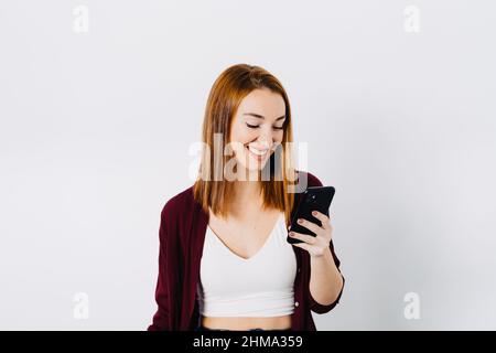 Cheerful young ginger haired female in top browsing mobile phone while standing against white wall and looking at screen Stock Photo