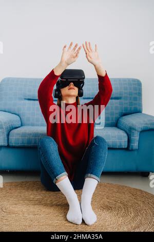Woman in VR goggles smiling with arms open and raised sitting against sofa and exploring virtual reality Stock Photo