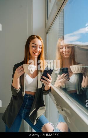 Happy red haired Woman standing against window while using mobile phone and drinking coffee on sunny day Stock Photo