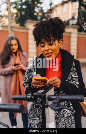 Young African American ethnic Woman with curly hair in warm coat standing on city street and doing contactless payment for electric scooter with smart Stock Photo