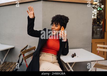 Excited curly haired African American female in trendy outfit sitting at table in outdoor cafe and waving hand while having phone conversation Stock Photo
