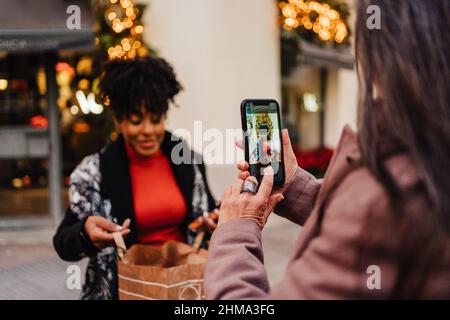 Selective focus of excited African American Woman demonstrating shopping bag to crop friend taking photo on smartphone on street Stock Photo