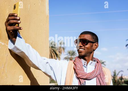 Positive Arab male in sunglasses taking self shot on mobile phone while standing in tropical park on sunny day Stock Photo