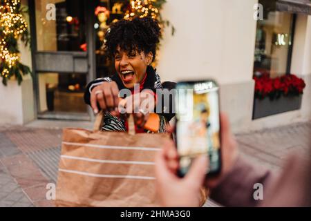 Selective focus of excited African American Woman demonstrating shopping bag to crop friend taking photo on smartphone on street Stock Photo