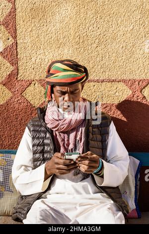 Serious Egyptian male wearing traditional turban and clothes surfing internet on smartphone while sitting on sofa in courtyard near house on sunny day Stock Photo