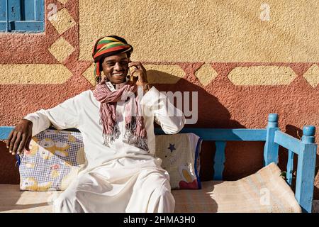 Cheerful Egyptian male wearing traditional turban and clothes talking smartphone while sitting on sofa in courtyard near house on sunny day Stock Photo