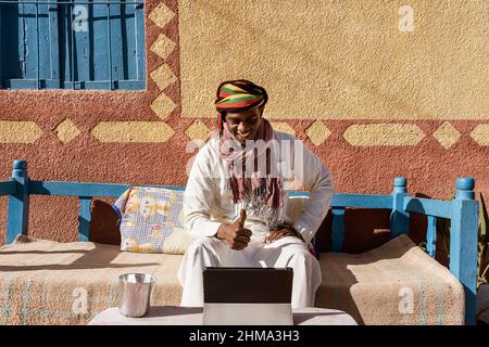 Sincere Egyptian male in traditional turban and robe having video chat on tablet while sitting on sofa in backyard on sunny day Stock Photo