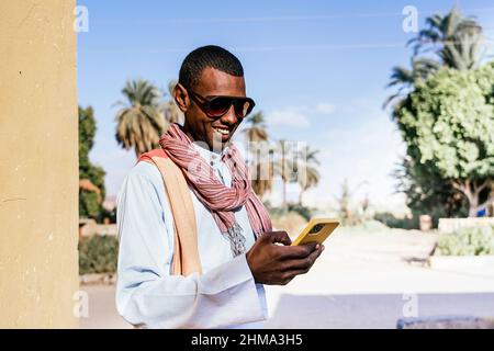 Smiling Arab male in traditional clothes and sunglasses messaging on mobile phone while standing in exotic park in summer Stock Photo
