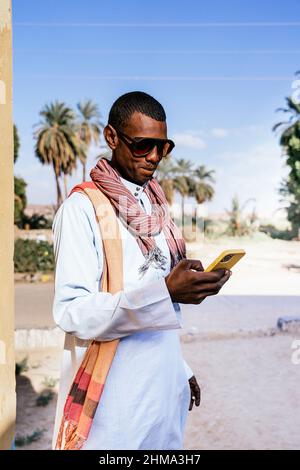 Smiling Arab male in traditional clothes and sunglasses messaging on mobile phone while standing in exotic park in summer Stock Photo