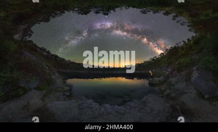 Side view of unrecognizable male tourist with torch admiring mounts reflecting in water under starry sky at night rocky coast with trees against dark Stock Photo