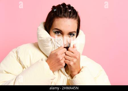 Attractive young female with makeup covering half of face with trendy warm white jacket and looking at camera on pink background Stock Photo