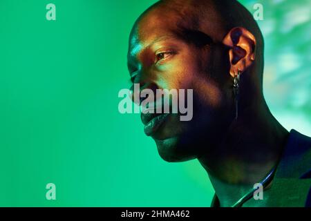 Confident bald African American male model with necklace and earring in trendy outfit standing at wall with glowing lights in modern studio looking aw Stock Photo