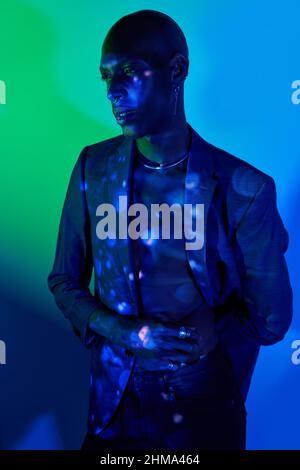 Serious bald African American slim male in necklace and stylish outfit standing with eyes closed in modern studio with dim illumination and glowing li Stock Photo