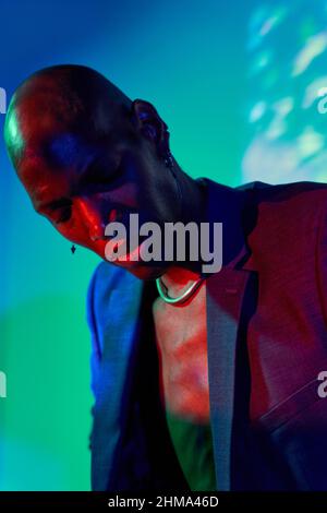 Confident bald African American male model with necklace and earring in trendy outfit standing with eyes closed at wall with glowing lights in modern Stock Photo