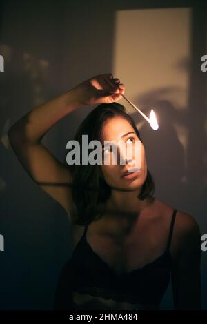 Young female with black hair looking at burning match in hand while standing at wall in room with dim light looking away Stock Photo