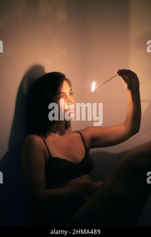 Young female with black hair looking at burning match in hand while standing at wall in room with dim light looking at fire Stock Photo