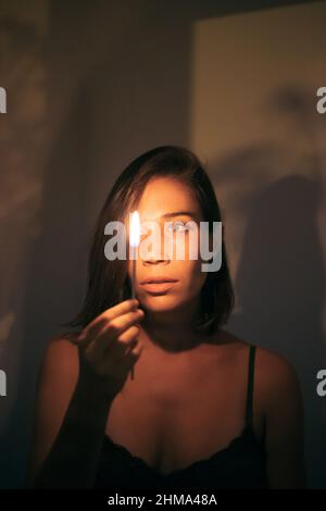 Young female with black hair looking at burning match in hand while standing at wall in room with dim light looking at fire Stock Photo
