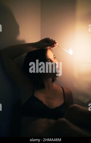 Young female with black hair looking at burning match in hand while standing at wall in room with dim light looking away Stock Photo