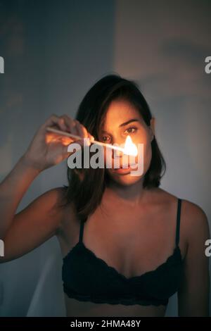 Young female with black hair looking at burning match in hand while standing at wall in room with dim light looking at fire Stock Photo
