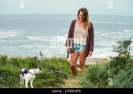 Optimistic female owner walking on grassy hill with obedient domestic cat on shore of foamy sea during in Cantabria Stock Photo
