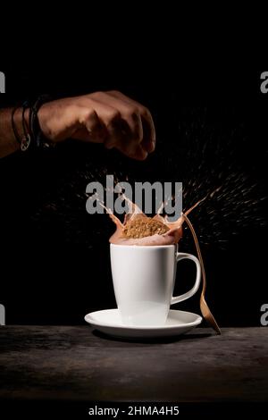 Crop anonymous person throwing tasty sweet cookies into white ceramic cup with splashing coffee on table in kitchen on black background Stock Photo