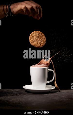 Crop anonymous person throwing tasty sweet cookies into white ceramic cup with splashing coffee on table in kitchen on black background Stock Photo