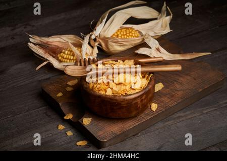 Bowl of tasty sweet cornflakes with spoon on wooden chopping board placed on table with corn cobs in light kitchen Stock Photo