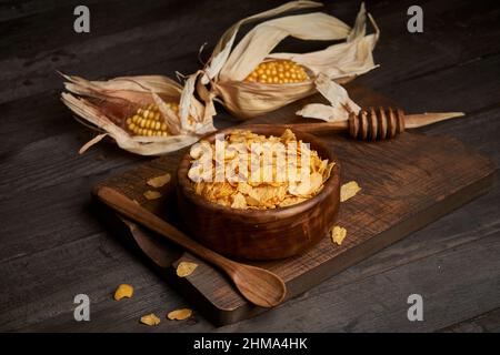 Bowl of tasty sweet cornflakes with spoon on wooden chopping board placed on table with corn cobs in light kitchen Stock Photo