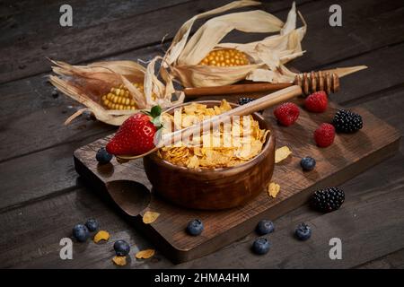 Bowl of tasty sweet cornflakes with spoon on wooden chopping board placed on table with corn cobs and berries in light kitchen Stock Photo