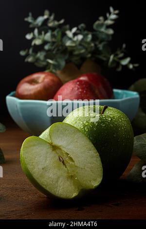 Wet green apple and a half placed on wooden table against blurred background of bowl with red apples near leaves Stock Photo